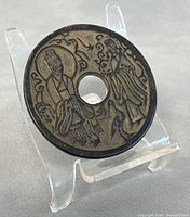 Photo of the bronze Fengshui coin on a clear stand showing the detailed engraving of two figures and ornamental design.