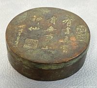 Photo of the round cast bronze trinket box top lid showing Chinese engraved characters and stamped seals in gold tone.