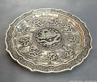 Top angled view of vintage white copper carved dragon plate showing raised dragon relief and scalloped rim detail.