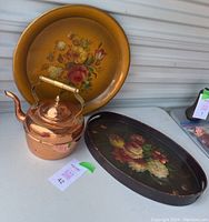 Photo showing all three items: round yellow floral tray, oval black floral tray, and polished copper kettle with brass handle.