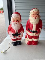Front view of two Santa Claus blow molds, untested, on a white surface outdoors. Standing side by side showing overall appearance, one shorter and one taller.