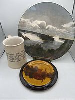 Three ceramic items including a scenic plate, text-inscribed mug, and a glazed trivet on display.