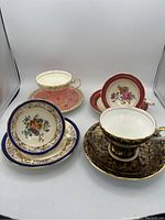 Four Aynsley teacups and matching saucers, each with distinct floral and decorative patterns, arranged on a white surface.