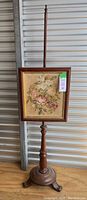 Full view of the antique fire screen mounted on a wooden stand with tripod base, featuring a framed needlepoint floral panel.