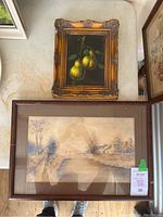 Two framed paintings on a table; one smaller ornate framed oil still life of pears and one larger watercolor landscape in simple frame.