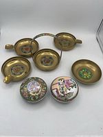 Full lot showing five brass ashtrays with colorful cloisonné-style enamel inserts and two round covered trinket dishes with painted Asian motifs.