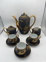Full view of tea set showing black porcelain with gold painted landscape scene including birds and trees. Set includes teapot, sugar bowl, creamer, and three cups and saucers.