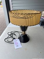 Photo of the entire lamp showing the beige fiberglass shade with black linear pattern, black metal base, and gold supporting rods. Power cord visible.