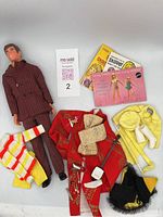 Full view showing the vintage Ken doll in plaid suit, assorted doll clothes including red kimono robe, yellow clothing, black and yellow hat, striped sweater, and small accessories. Also shows vintage Barbie and Skipper booklets.