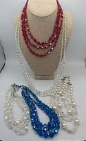Five vintage multi-strand necklaces arranged on a fabric bust and flat surface. Red, clear and white pearl-like necklaces visible.
