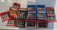 14 packages of mini glass Christmas ornaments spread on a flat surface showing various colors including metallic silver, gold, red, green and multi-colored sets, some original packaging with visible vintage designs.