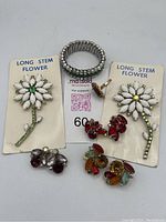 Photo showing two long stem flower pins on cards with white petals and green centers, silver-tone rhinestone stretch bracelet, and four pairs of rhinestone clip-on earrings in red, purple and multi-colored clusters.