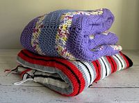 Two folded hand crocheted afghans stacked on a white surface, showing the blue/lavender multicolor and the red/grey/black/white striped patterns.