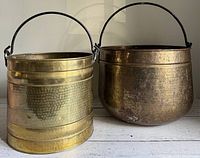 Two large vintage hammered brass planter pots placed side-by-side, showing their relative size, shape, and iron handles on a white surface.