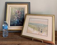 Two original framed paintings displayed side by side on a wooden surface with a water bottle for scale.