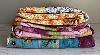 Stacked view of three colorful cotton quilts showing various batik and patchwork fabrics in bright colors including purple, turquoise, orange, and green