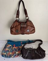 Three purses hanging and laid out showing overall appearance. Brown leather handbag, colorful tropical print crossbody bag, dark Kipling shoulder bag.