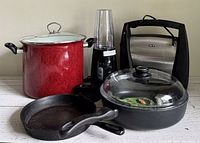 Full view of all 5 items in lot including stock pot, electric grinder, sausage grill, titanium skillet, and cast iron skillet.