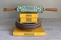 Stacked yellow Bistro Brie baker and under plate in box with green and white Anthropologie ceramic rolling pin on top and four stacked Manoir Core stoneware plates underneath