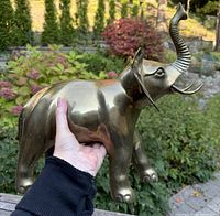Individual close-up showing detail and size of one brass elephant figure held in hand outdoors with foliage background.