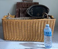 Front view of large rectangular wicker basket with handles, two flat twig trays and small oval vintage tray without insert visible, bottle of water for scale