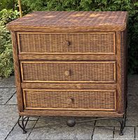 Photo of a wicker chest of drawers with three drawers and a metal base set on outdoor stone flooring with greenery background.