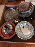 Box of assorted glass and metal ashtrays showing various shapes and stained conditions along with a metal ashtray with red finish.