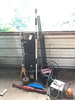 Wide view showing all items in lot leaning against garage wall: two metal jack stands, metal creeper, battery charger, broom, oil can, and shop light.