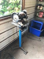 Full view of black bench grinder mounted on blue metal stand, positioned near window in workshop.