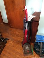 Red upright Bissell steam cleaner with hose and cord coiled around handle, placed on wooden floor near wooden door.