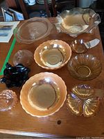 Photo shows nine assorted bowls on wooden table including peach lusterware carnival glass bowls, black nut dish, clear ribbed butter dish with lid, amber and clear depression glass bowls.