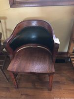 Front view of wooden arm chair showing seat, curved wood armrests, and black upholstered backrest with nailhead trim.