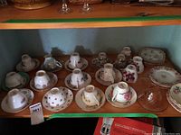 Multiple teacups and saucers arranged on a wooden shelf showing various floral designs and brands.