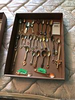 All collector spoons laid flat in cardboard box, showing variety of sizes and decorative handles.
