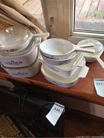 Seven CorningWare casserole dishes and bakeware in Blue Cornflower and Spice of Life patterns, some with glass lids, shown on a wooden surface indoors next to a window.