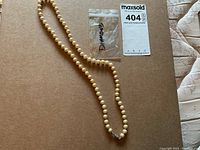 Faux pearl necklace laid out on a brown cardboard surface with a small plastic bag labeled Diamond containing two tiny loose diamonds and a MaxSold tag with lot number 404 visible.