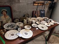 Wide shot of table displaying numerous commemorative plates, cups, tins, and a book related to British Royal Family figures.