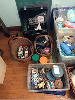Overall view of black Singer Featherweight machine case, wicker baskets and plastic containers of thread, yarn and notions on hardwood floor