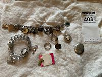 Overview of lot showing two charm bracelets, two rings, single earring, brooch, and locket pendant on fabric background.