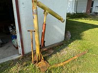 Full view of yellow hydraulic hoist standing upright on grass with visible rust and hydraulic piston.