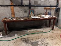 Wide view of the long wooden table against a concrete block wall with various items on top (items not included). Floor is stained concrete with a green hose on the floor.