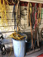 Wide view showing yellow extension cords hanging on wall and coiled in trash barrel with various tie-down straps and long-handled tools hung on the wall.