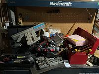 Photo of red metal toolbox and assorted tools including Benchmark Twister, cordless drill without charger, bits, hand tools, paint rollers, extension cords, carpenter's square, and miscellaneous hardware.
