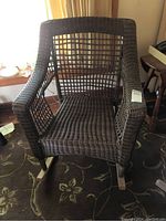 Full front view of the dark brown wicker rocking chair showing the woven seat, backrest, and arms, with wooden rockers on the bottom.