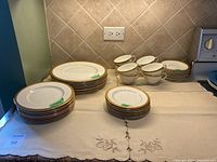 Set of gold-trimmed Royal Doulton china including stacks of dinner plates, side plates, dessert plates, cups, and saucers arranged on a countertop with linen placemats underneath.