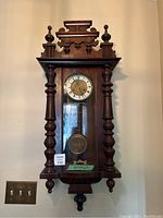 Full frontal photo of the antique German wall clock showing wooden case, clock face with Roman numerals, and visible pendulum behind glass door.
