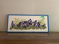 Front view of floral needlepoint art with purple flowers in blue frame
