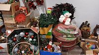 Photo showing boxes and bins containing multiple Christmas decorations including wreaths, string lights, figurines, and decorative mesh ribbons.