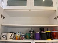 Photo showing ceramic coffee mugs, glass French press, and assorted insulated and plastic tumblers with straws arranged on a white shelf inside a cabinet.