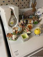Easter themed decorative items arranged on a white surface including ceramic bunnies, small flower pots, a yellow chick figurine, and a white egg-shaped container packed in Styrofoam.